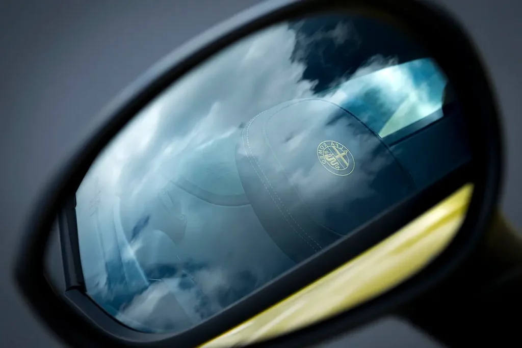 Vue artistique du rétroviseur d'une Alfa Romeo 4C Spider, révélant le logo Alfa sur l'appui-tête et le reflet des nuages.