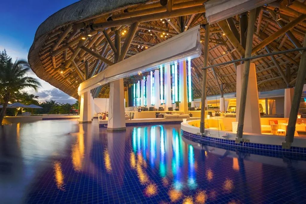 Vue nocturne du restaurant du So Sofitel Mauritius avec éclairages bleus se reflétant dans la piscine et structure du toit de chaume apparente.