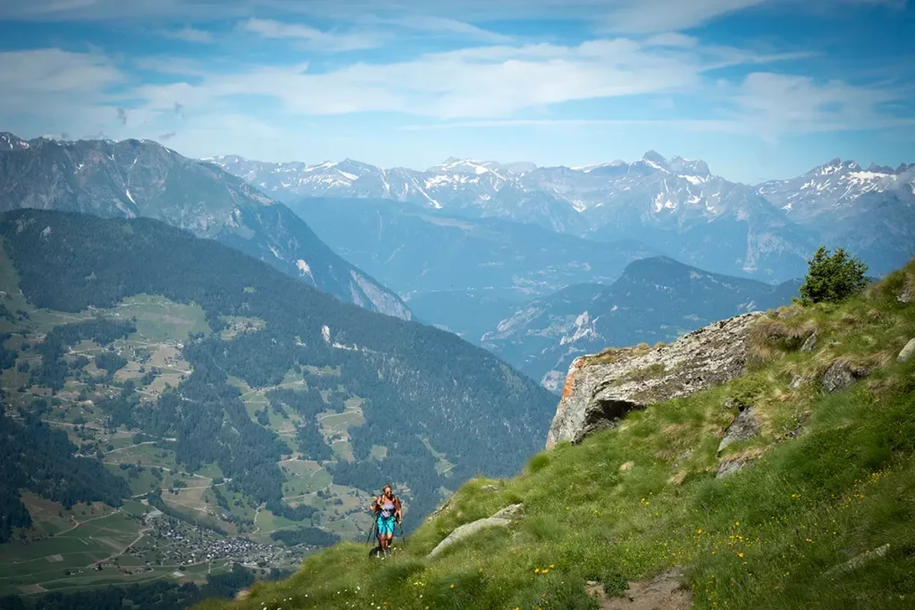 Photo en plongée d'un randonneur utilisant des bâtons pour monter vers le Col Termin à Verbier en été, avec vue sur la vallée.