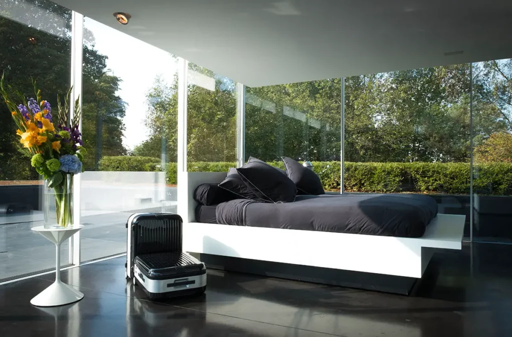 Chambre moderne et épurée de la Villa Panamera avec de grandes baies vitrées donnant sur la nature et des valises au pied du lit.