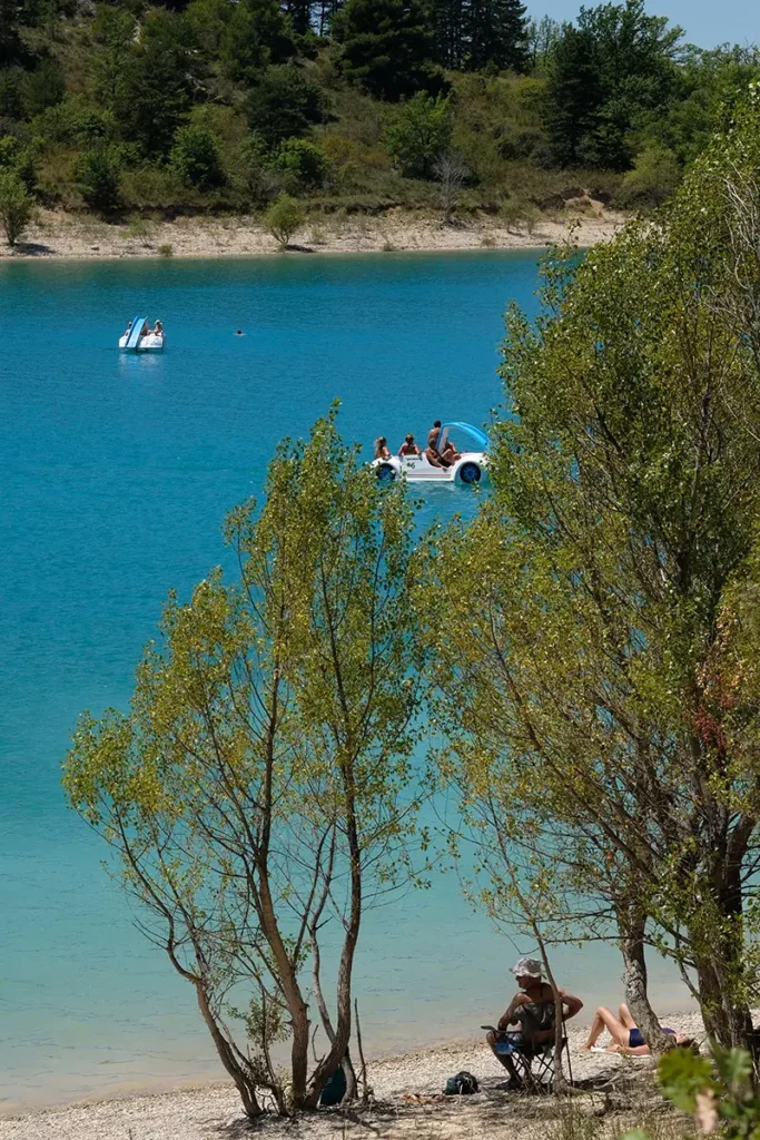 Photo verticale depuis la berge du lac de Sainte-Croix, montrant en avant-plan un homme au chapeau contemplant l'eau turquoise et deux pédalos, avec l'île de Costebelle au loin.