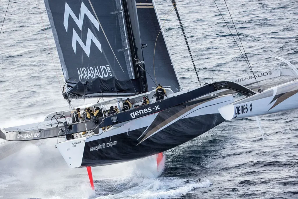 Vue trois-quarts arrière du trimaran Spindrift 2 sautant sur les vagues en mer houleuse, laissant apparaître ses foils orange fluo hors de l'eau.