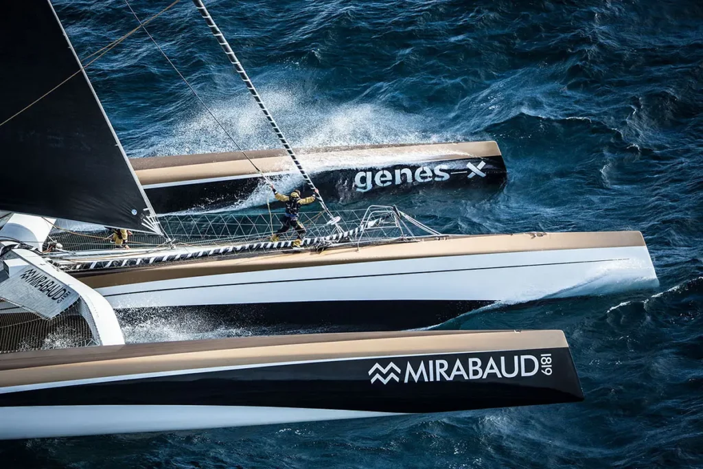 Gros plan sur la coque centrale et les flotteurs du trimaran Spindrift 2 frappant les vagues, avec un homme s'avançant prudemment en se tenant.