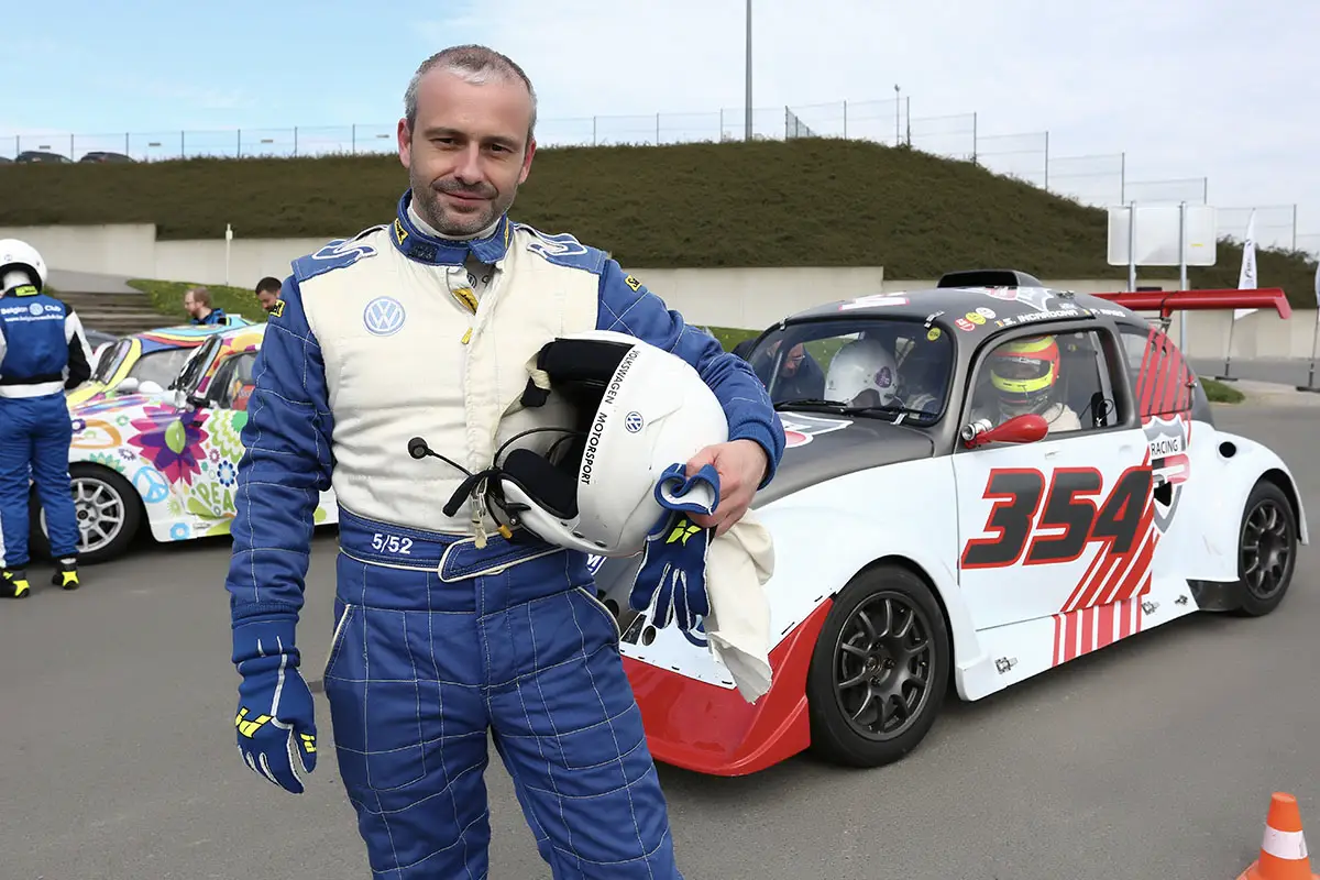 Le photographe et blogger Michel Gronemberger en combinaison de pilote bleue et blanche, casque sous le bras, sur le circuit de Mettet.