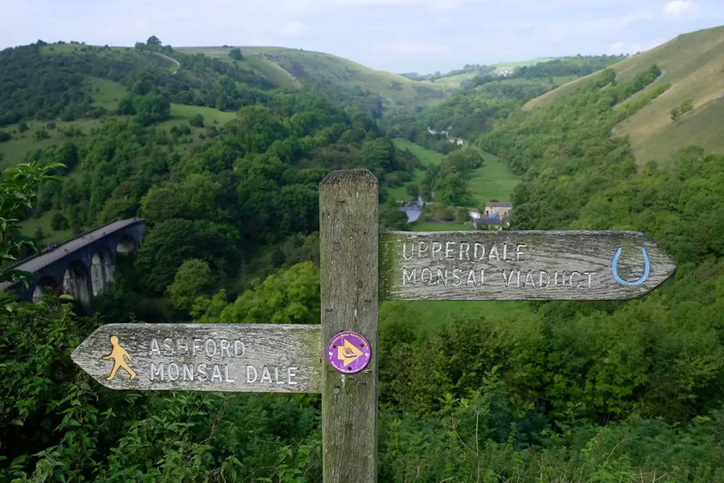 Panneau indicateur en bois à Monsal Head pointant vers Ashford, Monsal Dale et le viaduc, avec le pont ferroviaire en arrière-plan.