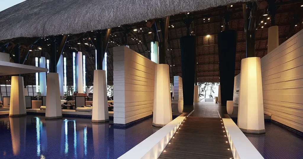 Passerelle du So Sofitel Mauritius avec colonnes en béton blanc, poutres en bois et toit de chaume sous un éclairage bleu moderne.