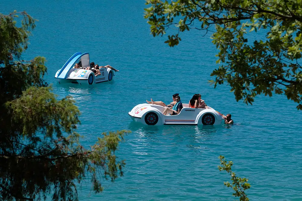 Deux pédalos en forme de voitures, dont un avec toboggan, sur les eaux calmes du lac de Sainte-Croix sous un soleil radieux.