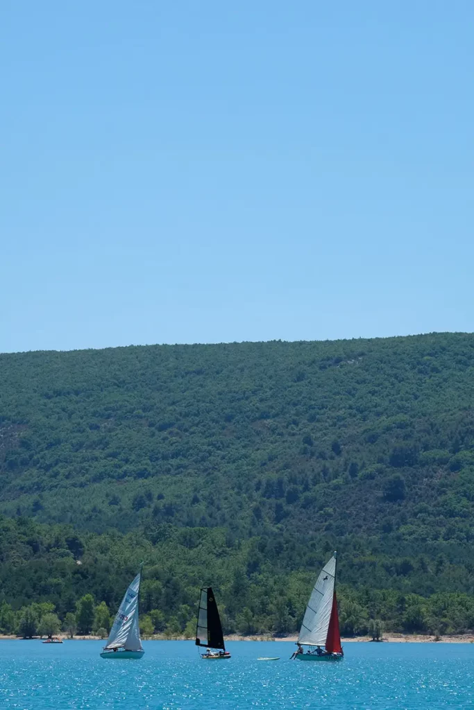 Vue verticale montrant trois petits voiliers naviguant sur les eaux turquoise du lac de Sainte-Croix, bordés par une végétation dense et un ciel bleu azur.