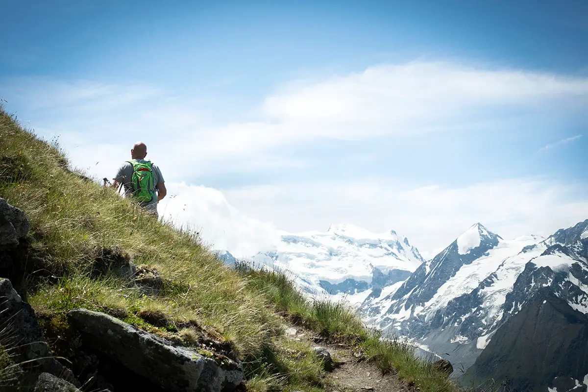 Randonneur de dos marchant sur le Sentier des Chamois avec une vue panoramique sur le massif des Combins à Verbier en été.