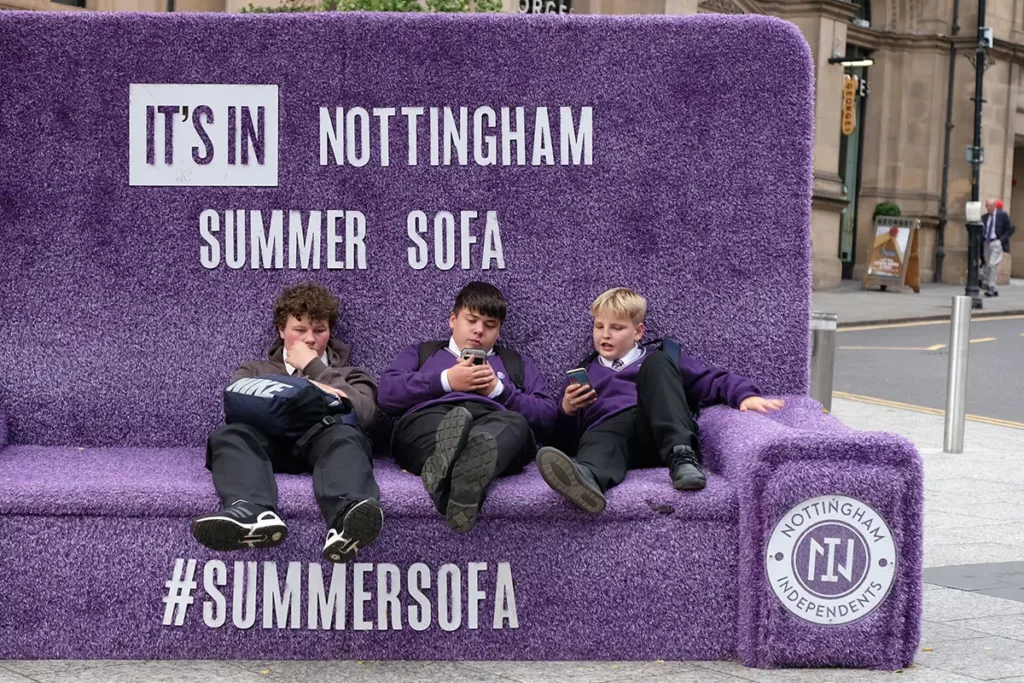 Trois écoliers en uniformes mauves allongés sur le Summer Sofa de Nottingham Independants, regardant leurs smartphones.