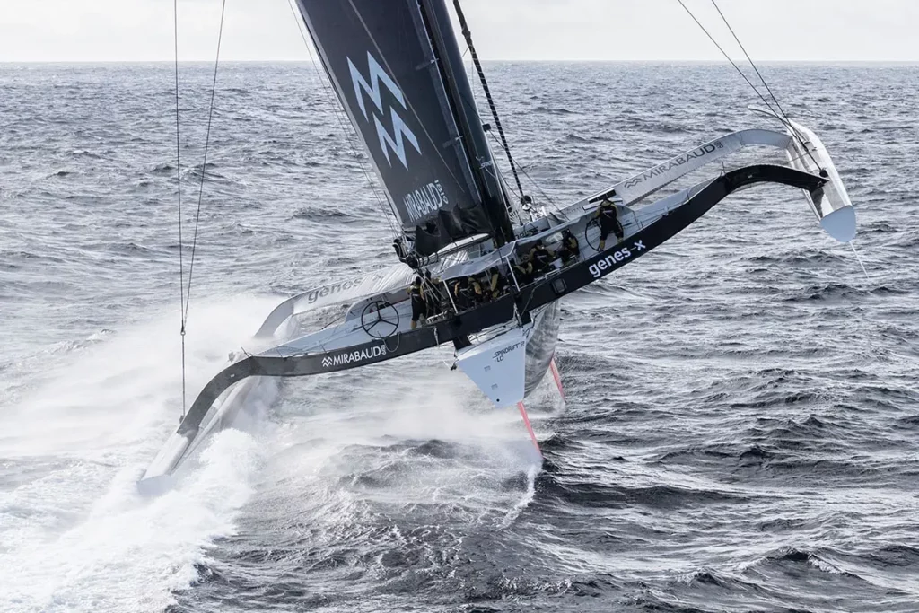 Vue aérienne arrière du trimaran Spindrift 2 avec un seul flotteur touchant l'eau, illustrant la puissance de la levée sous l'effet de la vitesse.