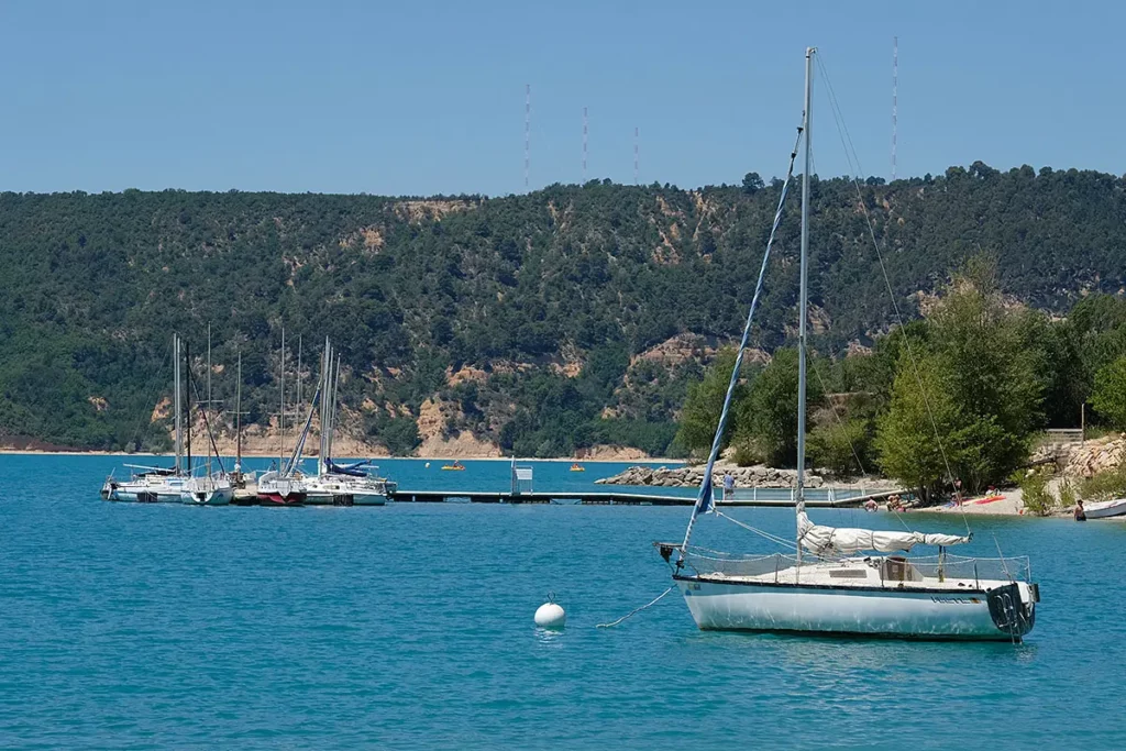 Photo horizontale montrant un voilier amarré en avant-plan sur le lac de Sainte-Croix, avec la plage des Salles-sur-Verdon et son ponton en arrière-plan.