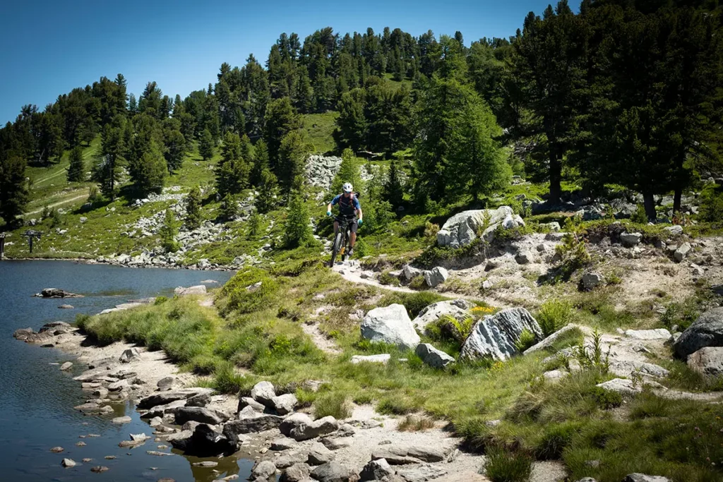 Cycliste en VTT électrique roulant sur un sentier bucolique longeant un lac de haute montagne sur le Tour du Mont-Fort à Verbier en été.