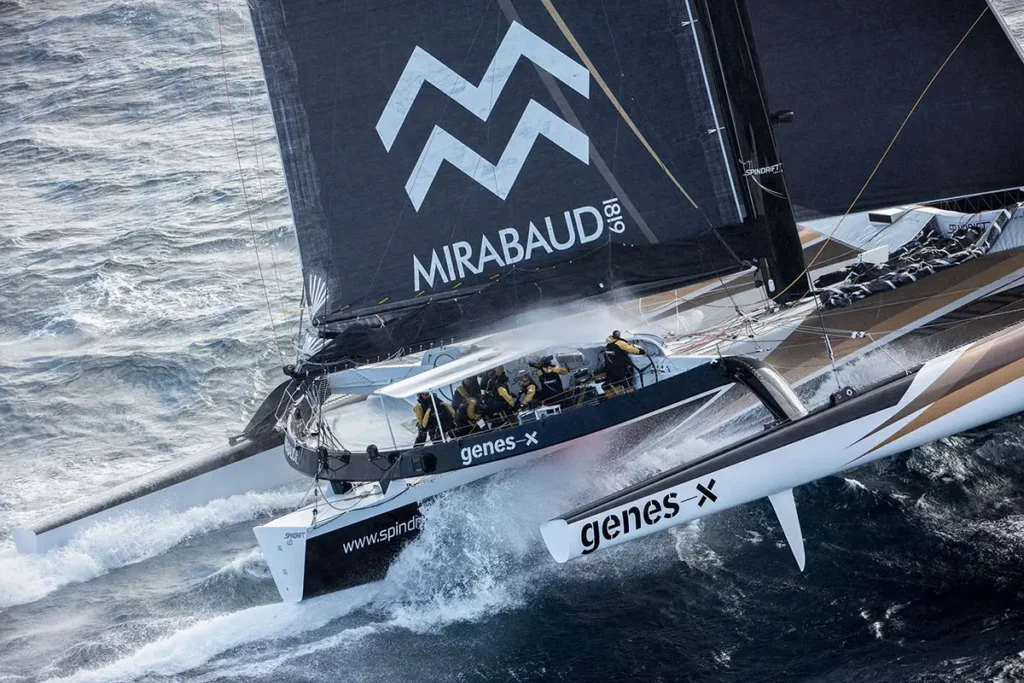 Vue trois-quarts arrière du cockpit de Spindrift 2 montrant le skipper Yann Guichard à la barre et son équipage en pleine manœuvre par mer houleuse.