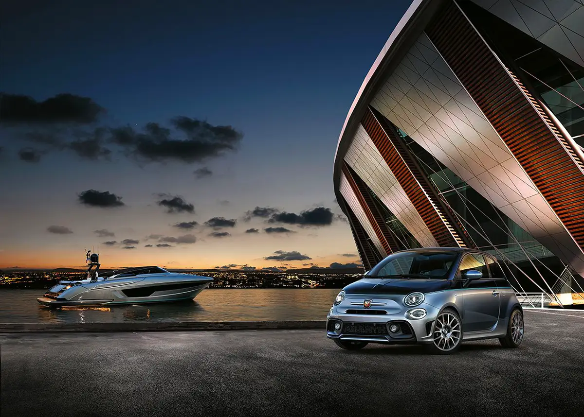 Abarth 695 Rivale stationnée sur un quai à côté d'un bateau Riva moderne et sportif sur un plan d'eau au crépuscule.