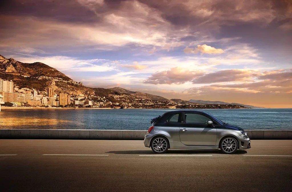 Vue de profil d'une Abarth 695 Rivale à l'arrêt sur une route de bord de mer avec la côte méditerranéenne illuminée en arrière-plan au crépuscule.