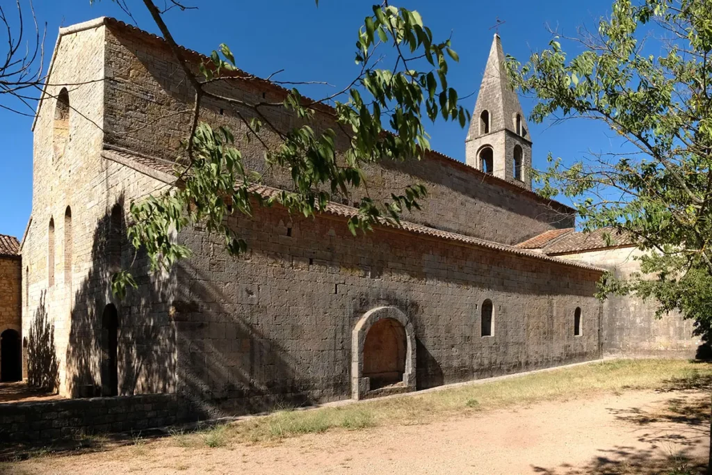 Vue panoramique de l'abbatiale de l'Abbaye du Thoronet montrant le pignon ouest ensoleillé, la façade sud avec l'enfeu et le clocher en arrière-plan.