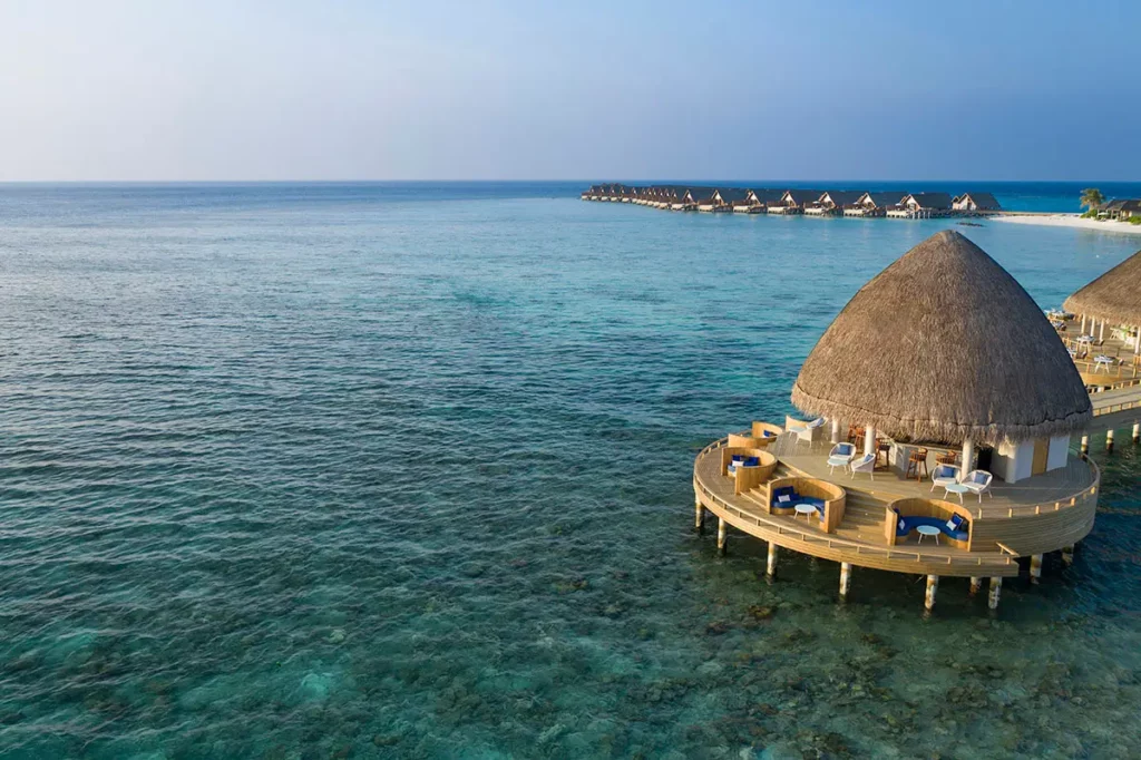 Vue aérienne du restaurant Boli à Emerald Faarufushi mettant en avant sa structure circulaire sur pilotis avec les villas sur l'eau en arrière-plan au milieu de l'Océan Indien.