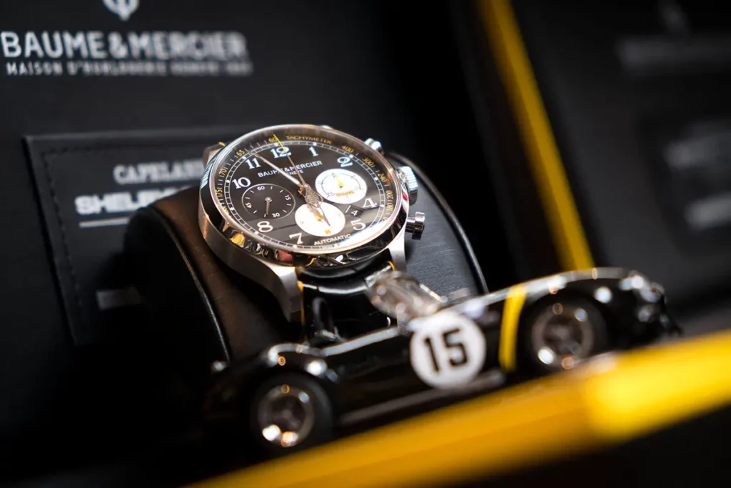 Photo horizontale du chronographe Baume & Mercier Capeland Shelby Cobra Classic dans son écrin, avec une voiture miniature Shelby Cobra noire numéro 15 en avant-plan floue.