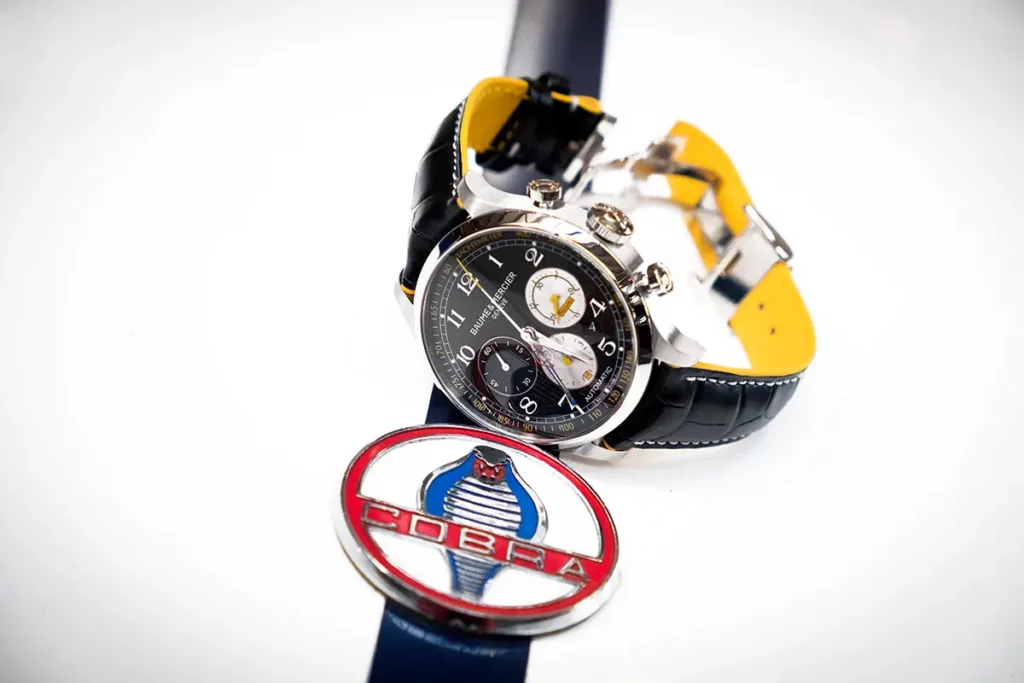 Gros plan du chronographe Baume & Mercier Capeland Shelby Cobra 1963 (modèle classique acier) posé sur le capot d'une voiture Shelby Cobra blanche à bande noire, juste au-dessus du logo Cobra.