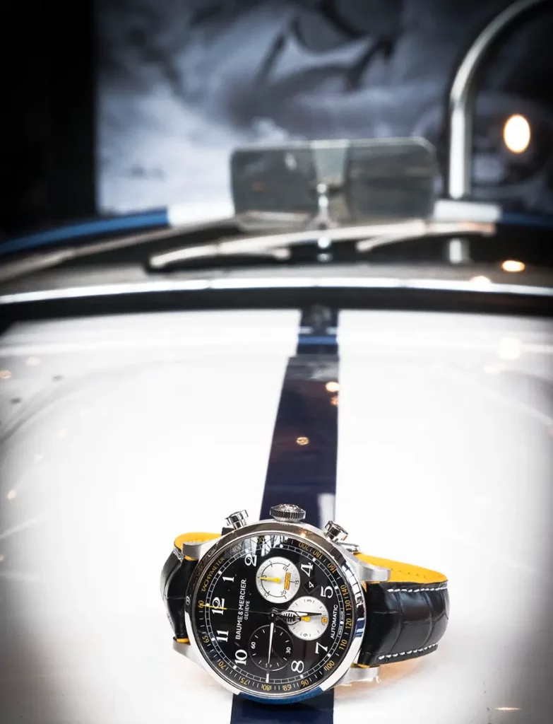 Photo verticale du chronographe Baume & Mercier Capeland Shelby Cobra posé sur la bande noire centrale d'un capot de voiture Shelby Cobra, avec le rétroviseur chromé visible dans le flou en arrière-plan.