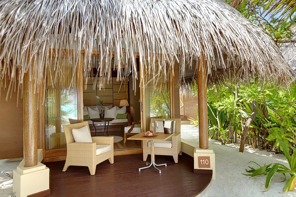 Entrée d'une Beach Villa au Kanuhura avec toit en chaume traditionnel, sable blanc et végétation luxuriante.