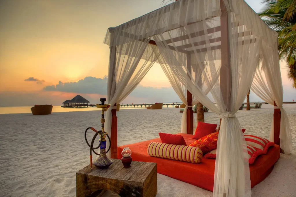 Lit à baldaquin orange sur la plage de sable blanc au Handhuvaru Bar du Six Senses Kanuhura lors d'un coucher de soleil aux Maldives.