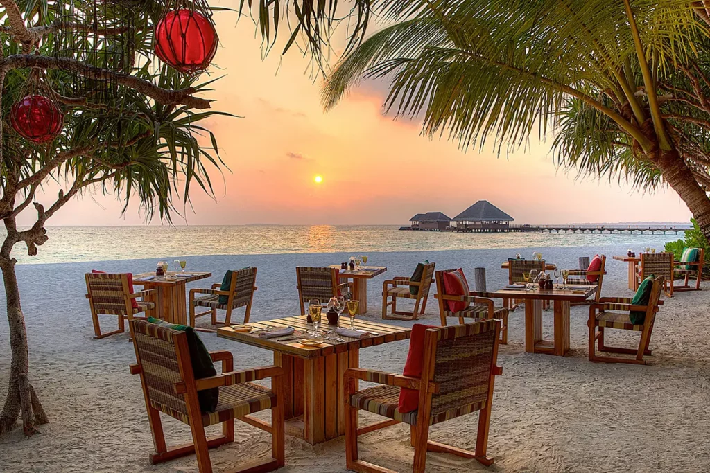Tables dressées sur le sable sous les palmiers au restaurant Thin Rah du Six Senses Kanuhura lors d'un coucher de soleil orangé.