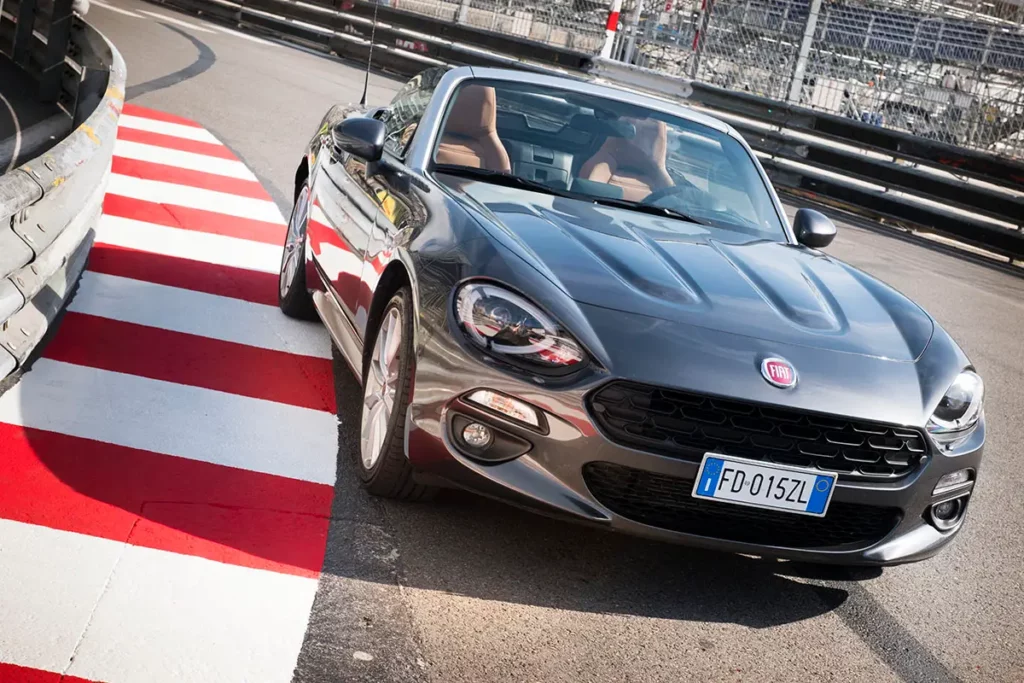 Fiat 124 Spider grise stationnée sur le circuit de Formule 1 de Monaco près des vibreurs rouges et blancs avant le Grand Prix.