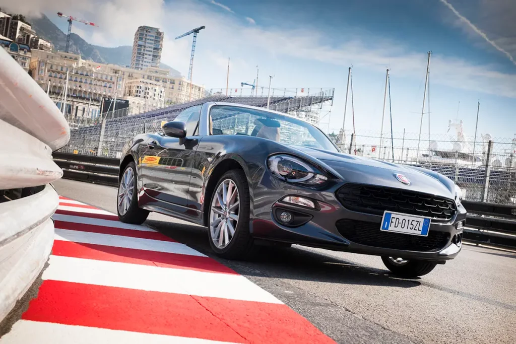 Vue en contre-plongée de la Fiat 124 Spider sur le circuit de Monaco avec vibreurs rouges, rails de sécurité, tribunes et mâts de bateaux dans le port.