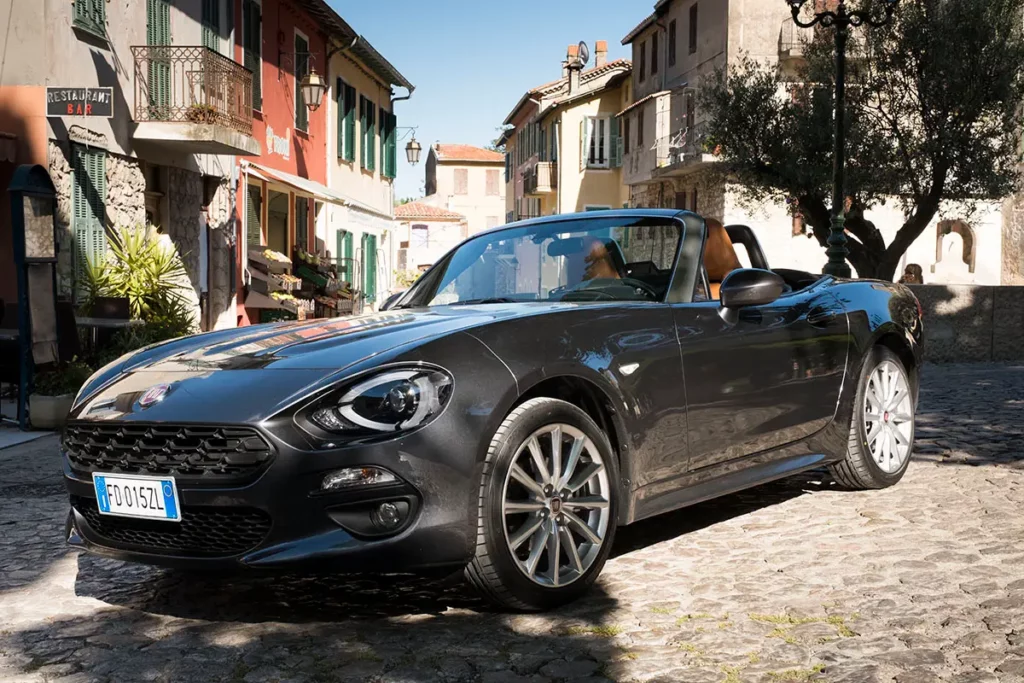 Prise de vue au niveau du sol en contre-plongée d'une Fiat 124 Spider sur la place pavée de Gorbio montrant l'épicerie et le bar-tabac.