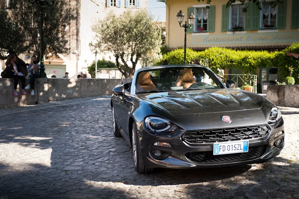 Vue trois-quarts avant d'une Fiat 124 Spider grise décapotée avec intérieur en cuir beige sous un platane sur la place de Gorbio.