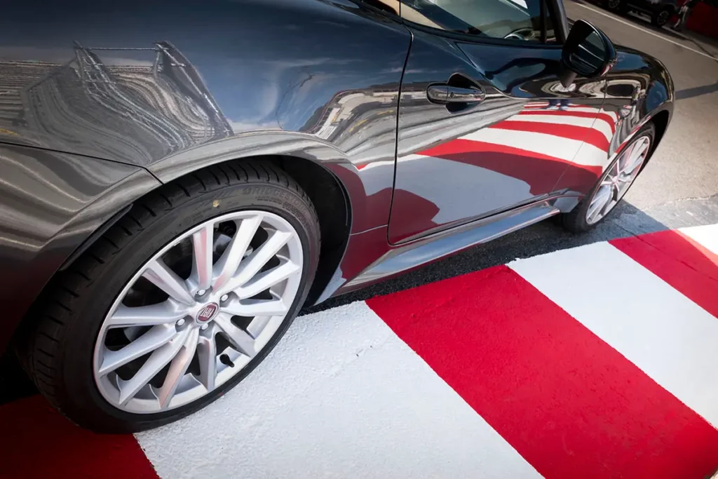 Gros plan sur le flanc de la carrosserie d'une Fiat 124 Spider grise reflétant les lignes rouges et blanches d'un vibreur du circuit de Monaco.