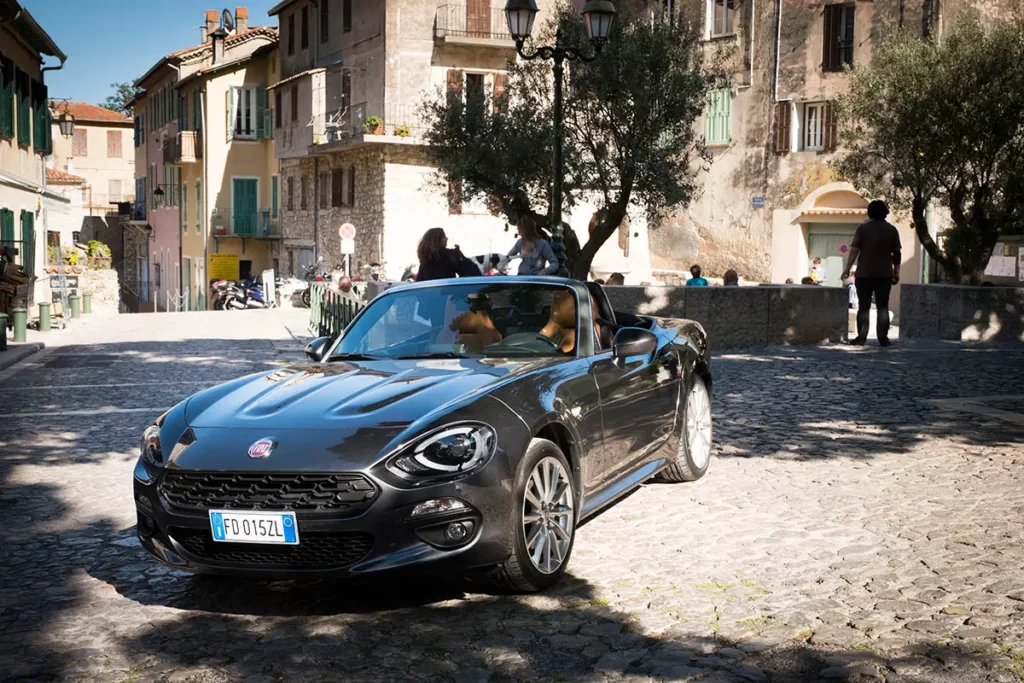 Vue de profil trois-quarts d'une Fiat 124 Spider aux reflets ensoleillés sur une place de Gorbio avec une perspective sur une rue provençale typique.