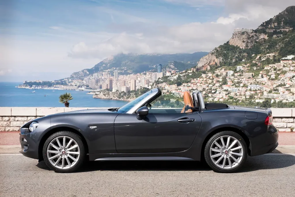 Vue latérale d'une Fiat 124 Spider grise décapotée sur une route de corniche surplombant la principauté de Monaco et les falaises de la Méditerranée.