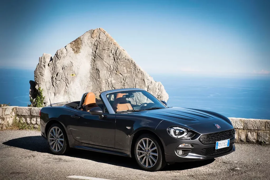 Fiat 124 Spider décapotée stationnée sur une aire de repos en montagne avec un muret de pierre et un rocher en forme de pyramide.