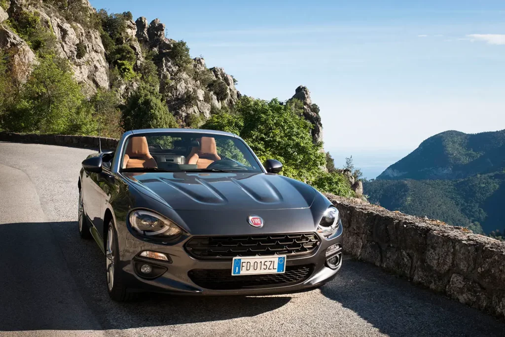 Vue de face d'une Fiat 124 Spider garée en pente à Sainte-Agnès avec un muret de protection, les montagnes de l'arrière-pays et la mer Méditerranée au loin.