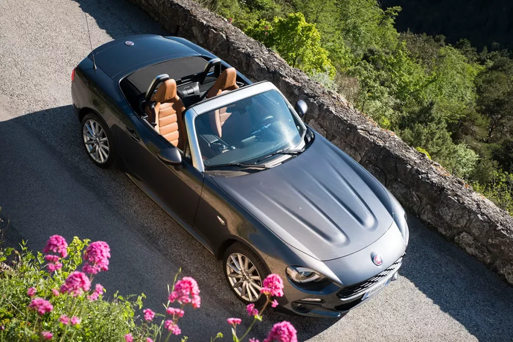 Vue en plongée d'une Fiat 124 Spider sur une route de montagne à Sainte-Agnès avec des fleurs roses au premier plan et le précipice en arrière-plan.