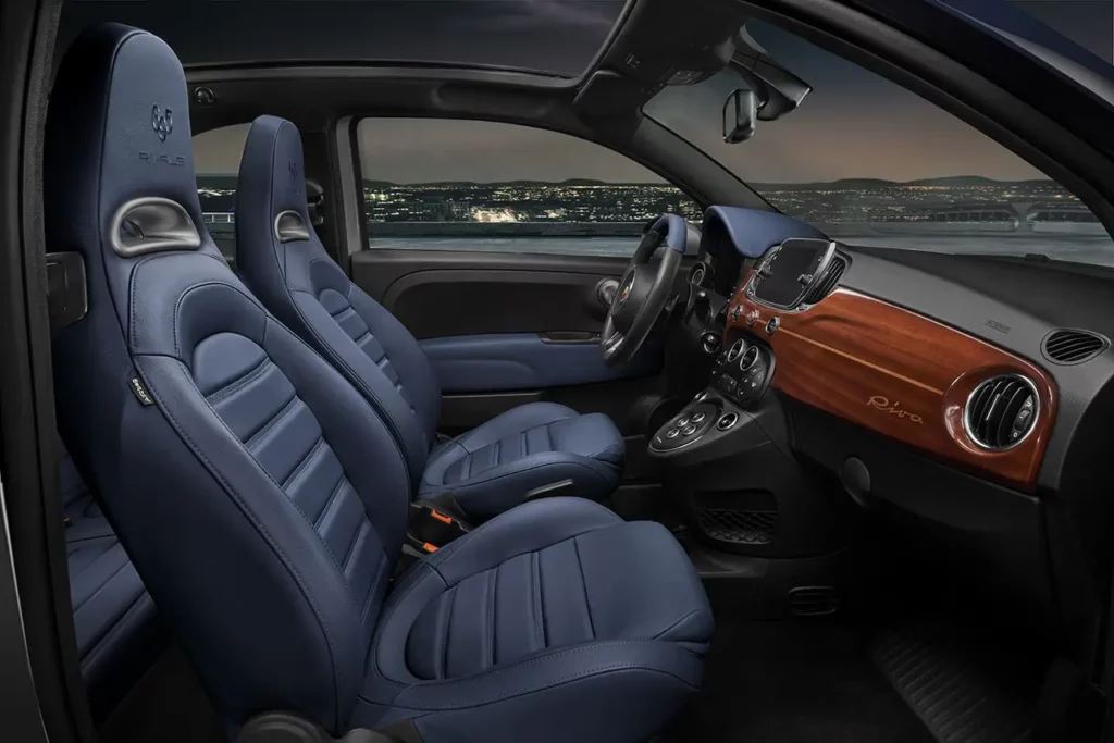 Vue de l'habitacle de l'Abarth 695 Rivale depuis la portière passager montrant les sièges baquets en cuir bleu et le tableau de bord en acajou Riva au crépuscule.