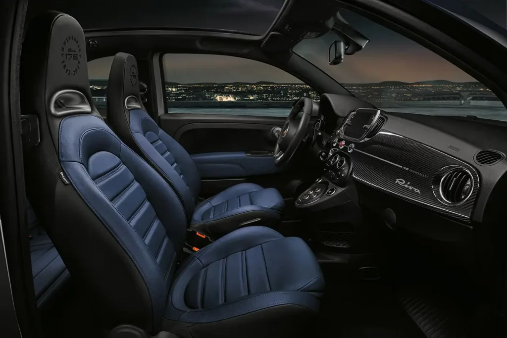 Vue de l'habitacle de l'Abarth 695 Rivale depuis le siège passager montrant le tableau de bord en carbone sous une lumière de fin de journée au bord de la mer.