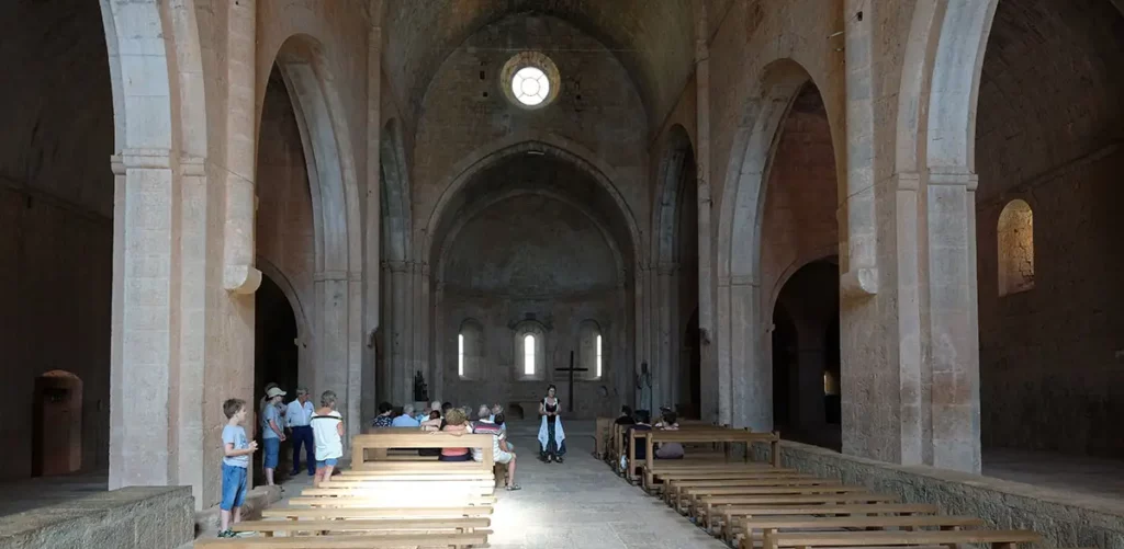 Vue intérieure de l'abbatiale du Thoronet depuis l'entrée ouest vers le chœur, montrant l'enfilade des bancs et un guide effectuant une démonstration d'acoustique.