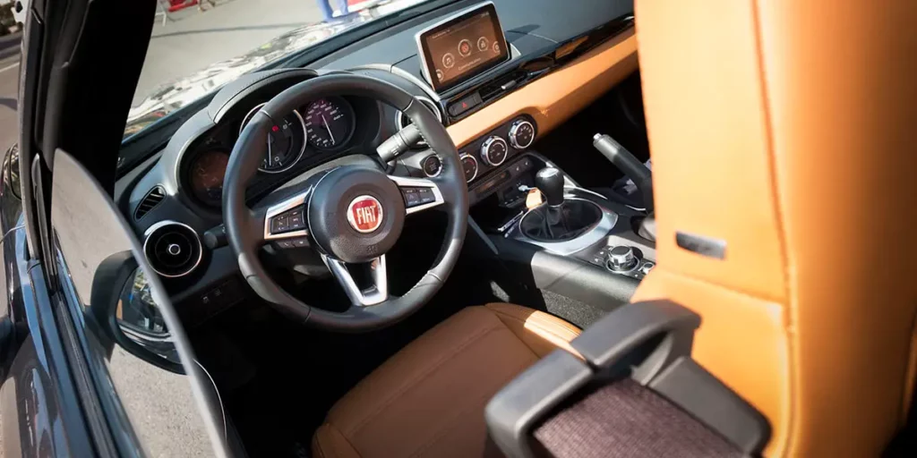 Vue de l'intérieur d'une Fiat 124 Spider avec volant en cuir, logo Fiat, console centrale avec navigation et levier de vitesse manuel.