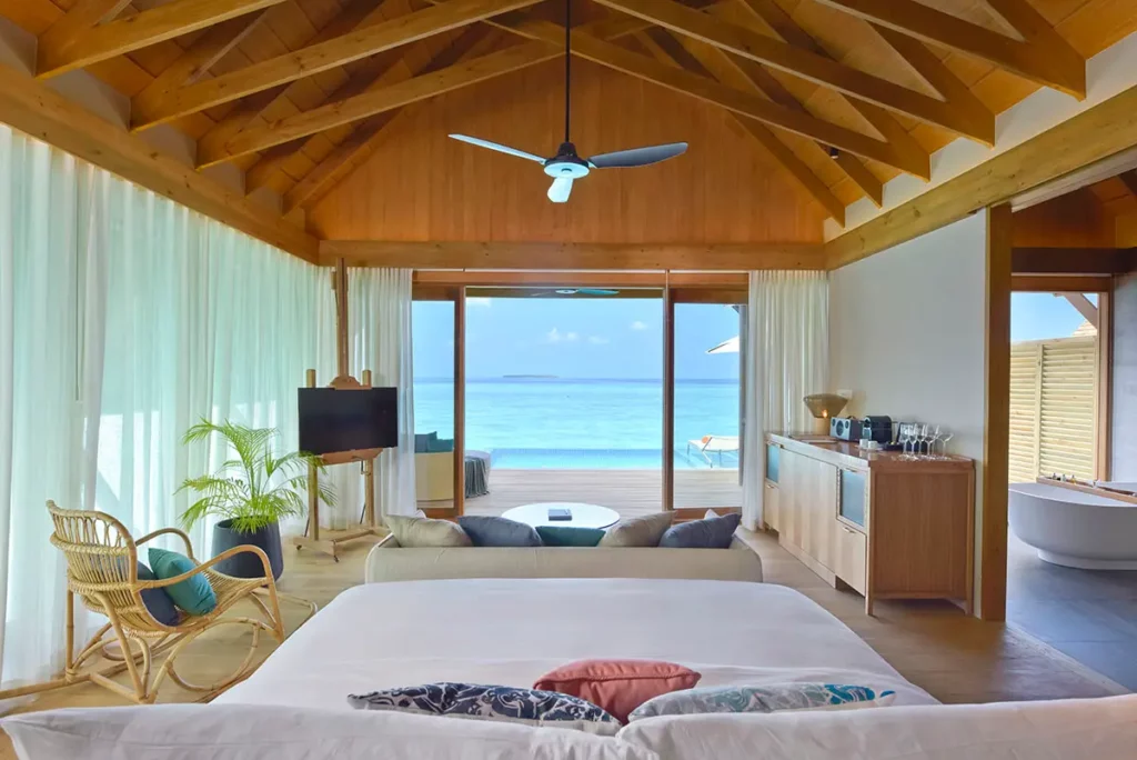 Vue depuis la tête de lit d'une Water Villa à Emerald Faarufushi, montrant l'enfilade vers le salon, la salle de bain et les baies vitrées ouvrant sur la piscine et l'Océan Indien.