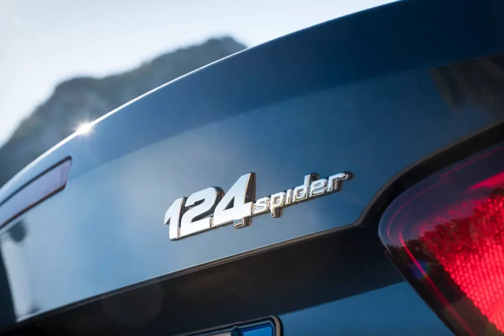 Gros plan horizontal sur le logo chromé 124 Spider sur le coffre de la voiture avec un reflet brillant du soleil.