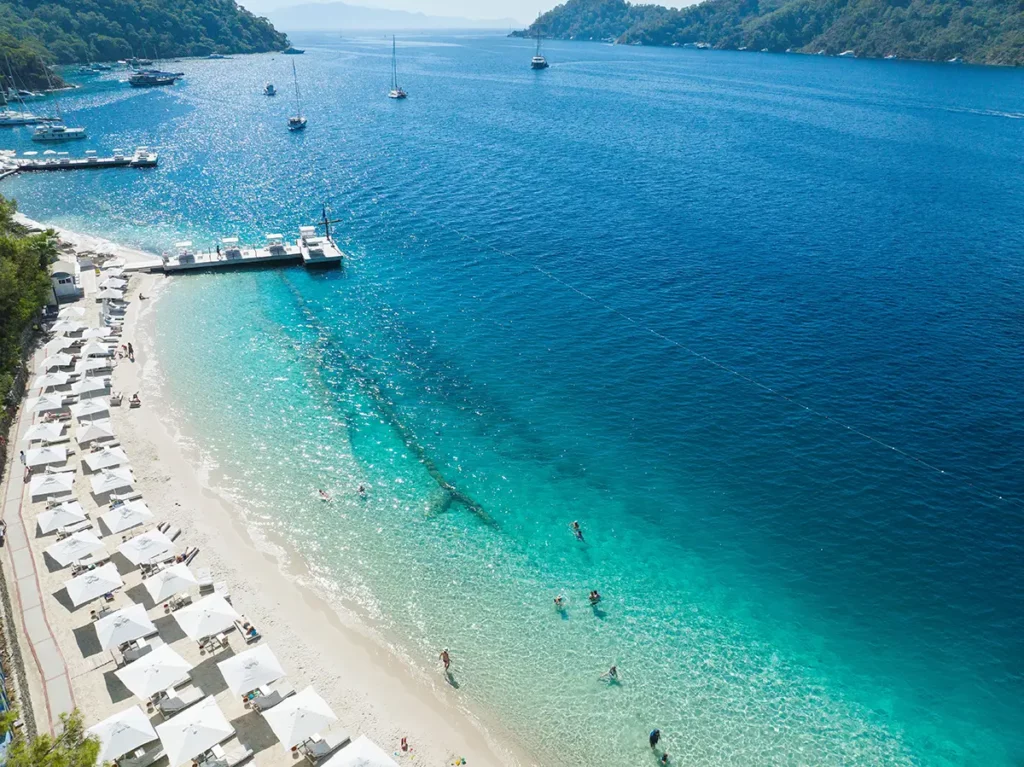 Alignement de parasols blancs sur la plage d'Ölüdeniz en Turquie, bordant une eau transparente qui vire au turquoise puis au bleu profond face aux collines.