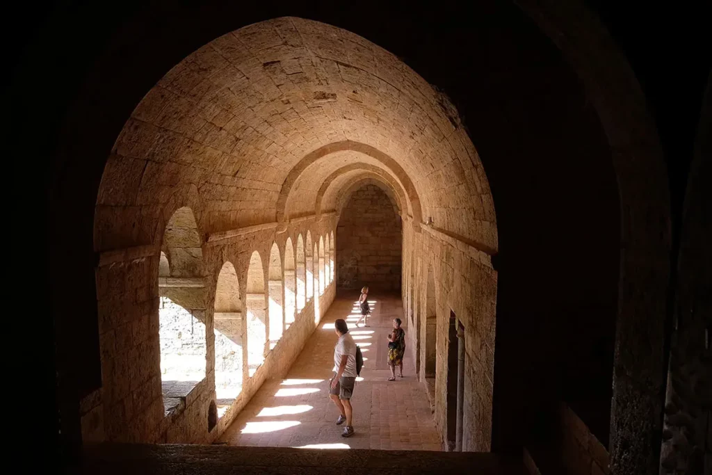 ue en perspective du couloir est du cloître de l'Abbaye du Thoronet, montrant les voûtes en pierre, les alcôves ensoleillées et des visiteurs.