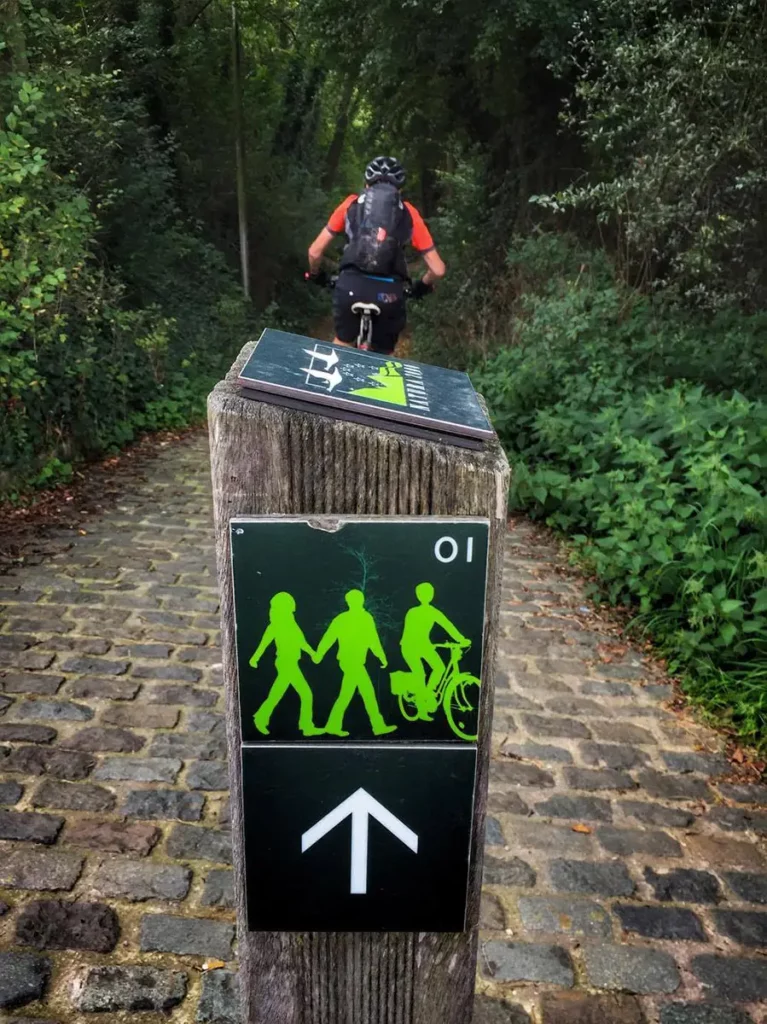 Poteau en bois de balisage de la Promenade Verte avec son logo vert reconnaissable, sur un tronçon pavé avec un cycliste de dos.