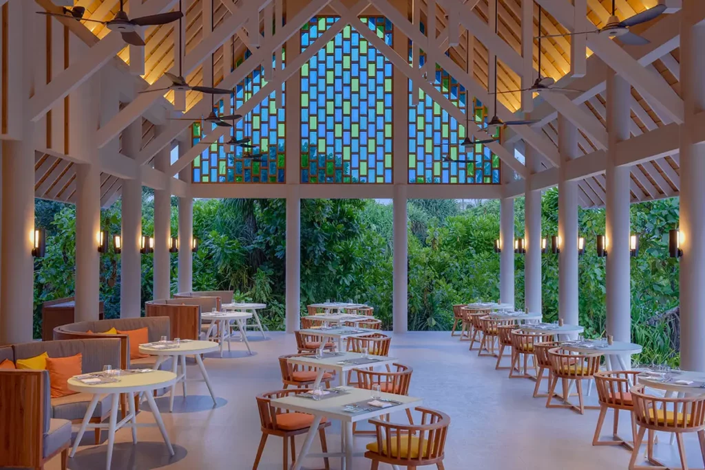 Vue de l'enfilade de tables du restaurant Iru à Emerald Faarufushi, mettant en avant sa structure aérée rythmée par une charpente et des piliers en bois.