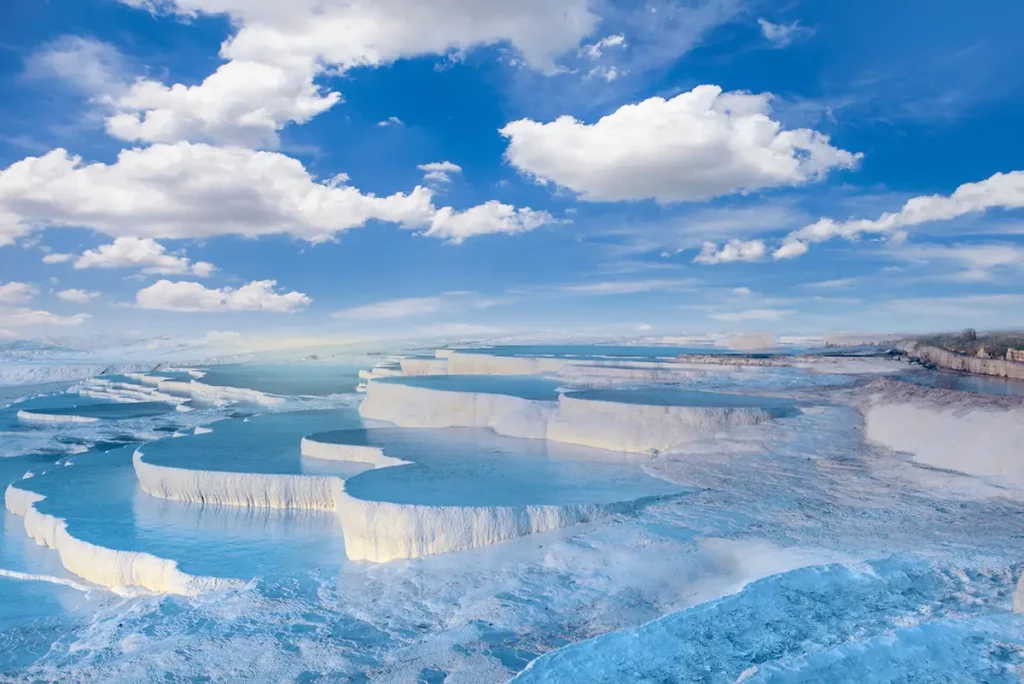 Vasques de travertin blanc de Pamukkale en Turquie, avec leurs piscines naturelles d'eau turquoise en escalier sous un ciel bleu clair parsemé de nuages.