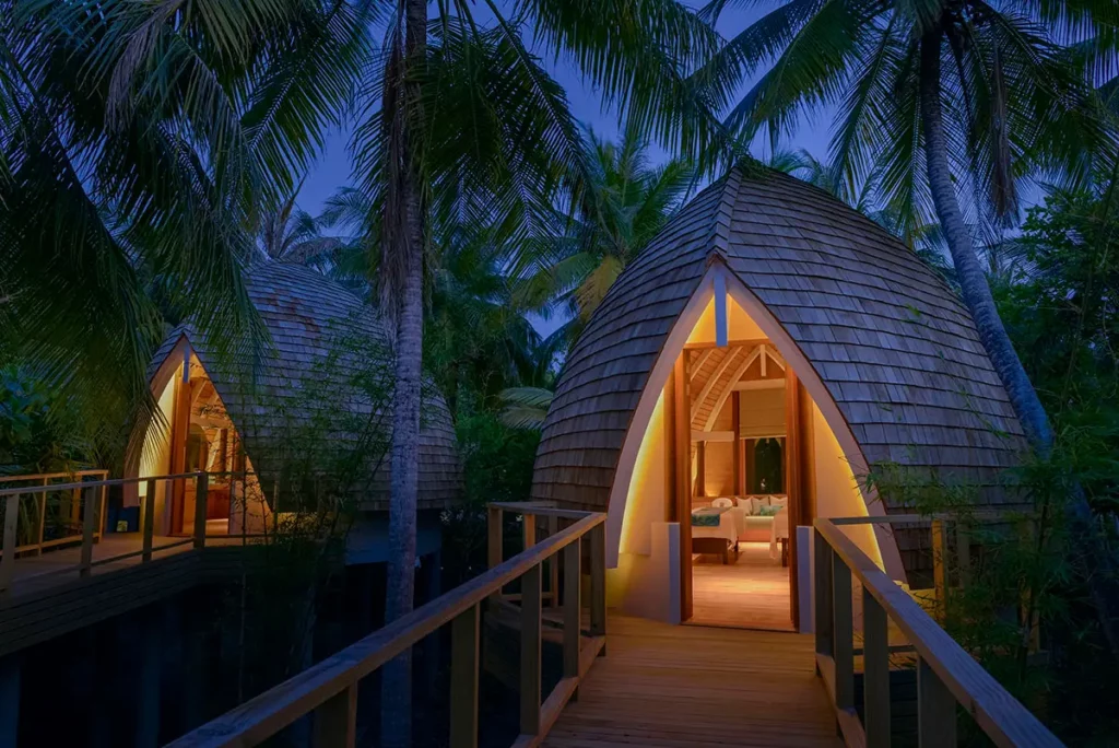 Vue nocturne de deux cabines sur pilotis du Spa Nika à Emerald Faarufushi, avec éclairage intérieur chaleureux sous un ciel bleu profond.